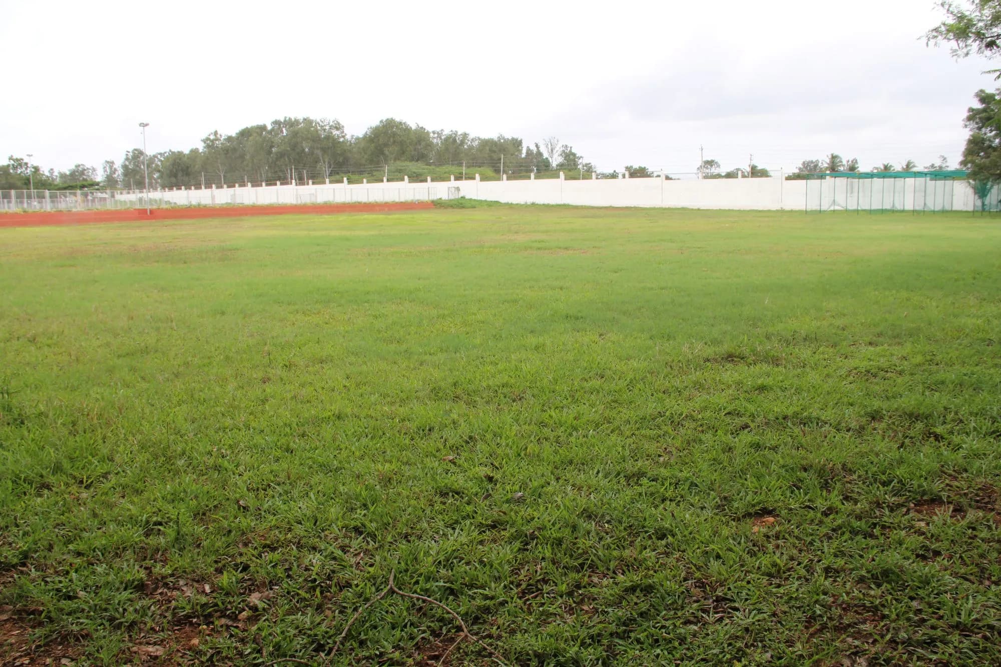 Turf-level view across the grass of the PSSEMR football ground, with the athletics track and boundary wall behind.