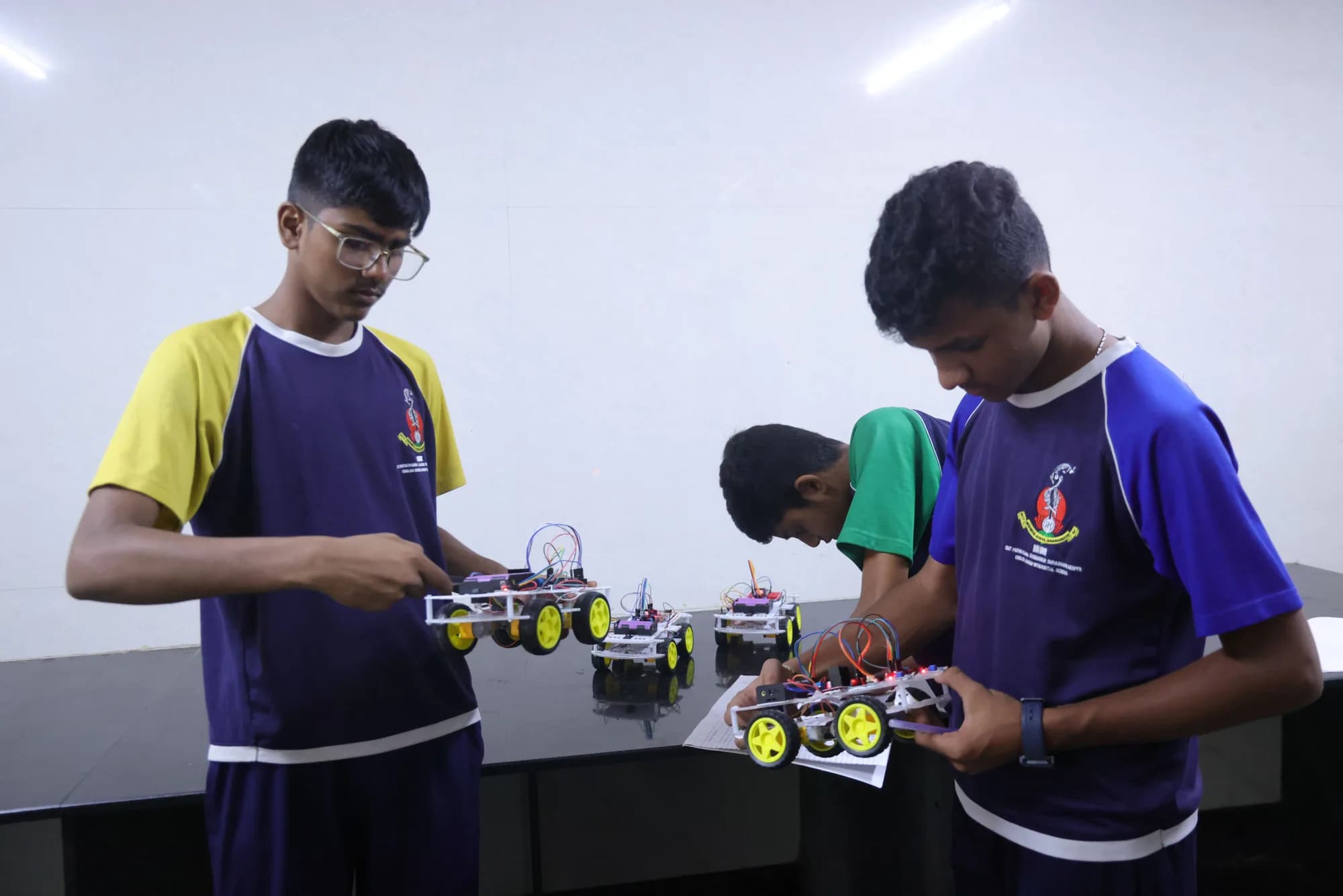Students in three different house shirts inspecting four-wheel robot builds