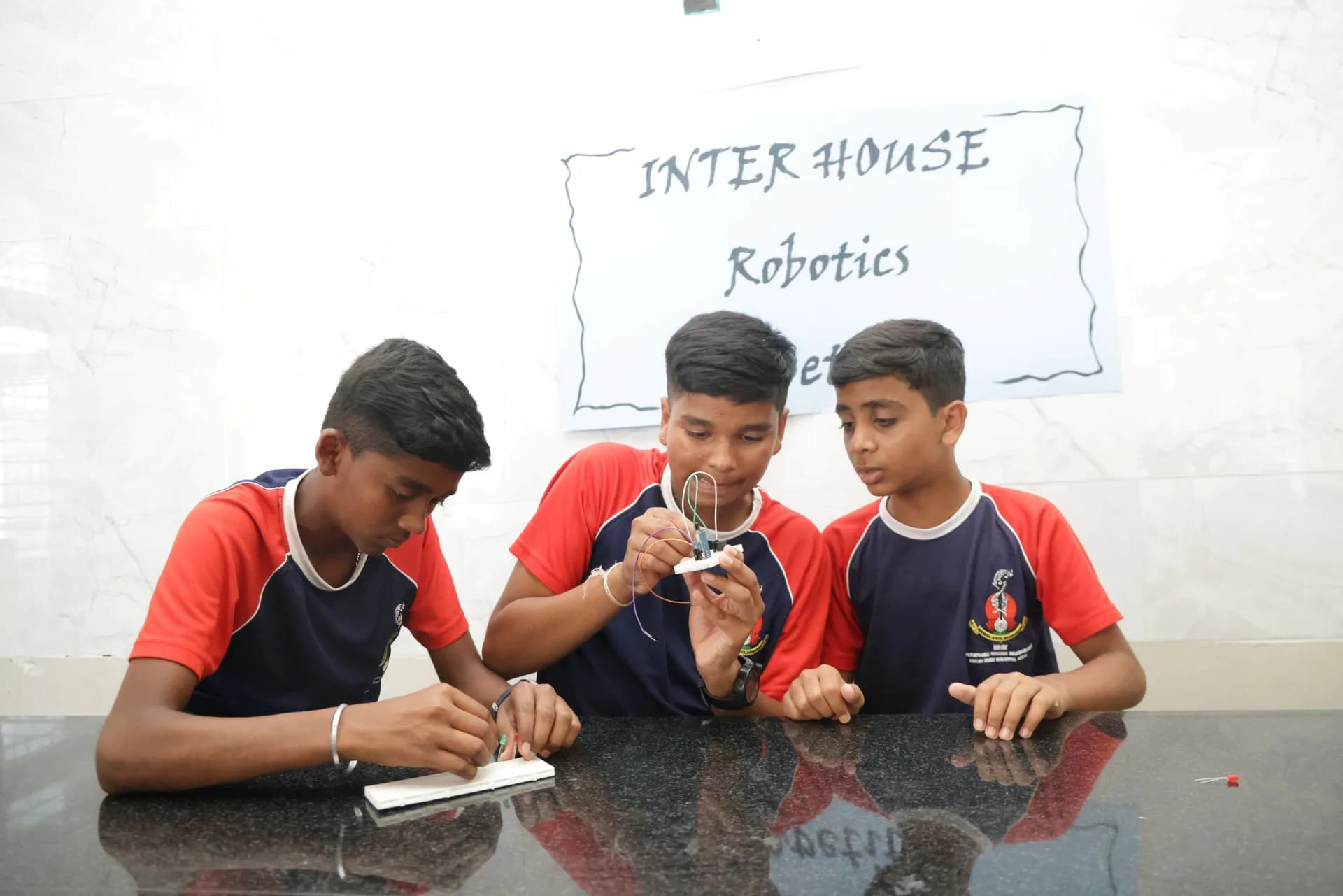 Three boys wiring a breadboard circuit at PSSEMR School, beneath a handwritten robotics sign