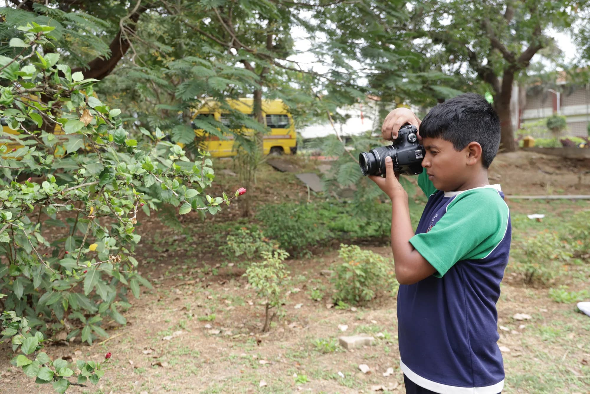 A PSSEMR School student framing a hibiscus flower through a DSLR, buses behind