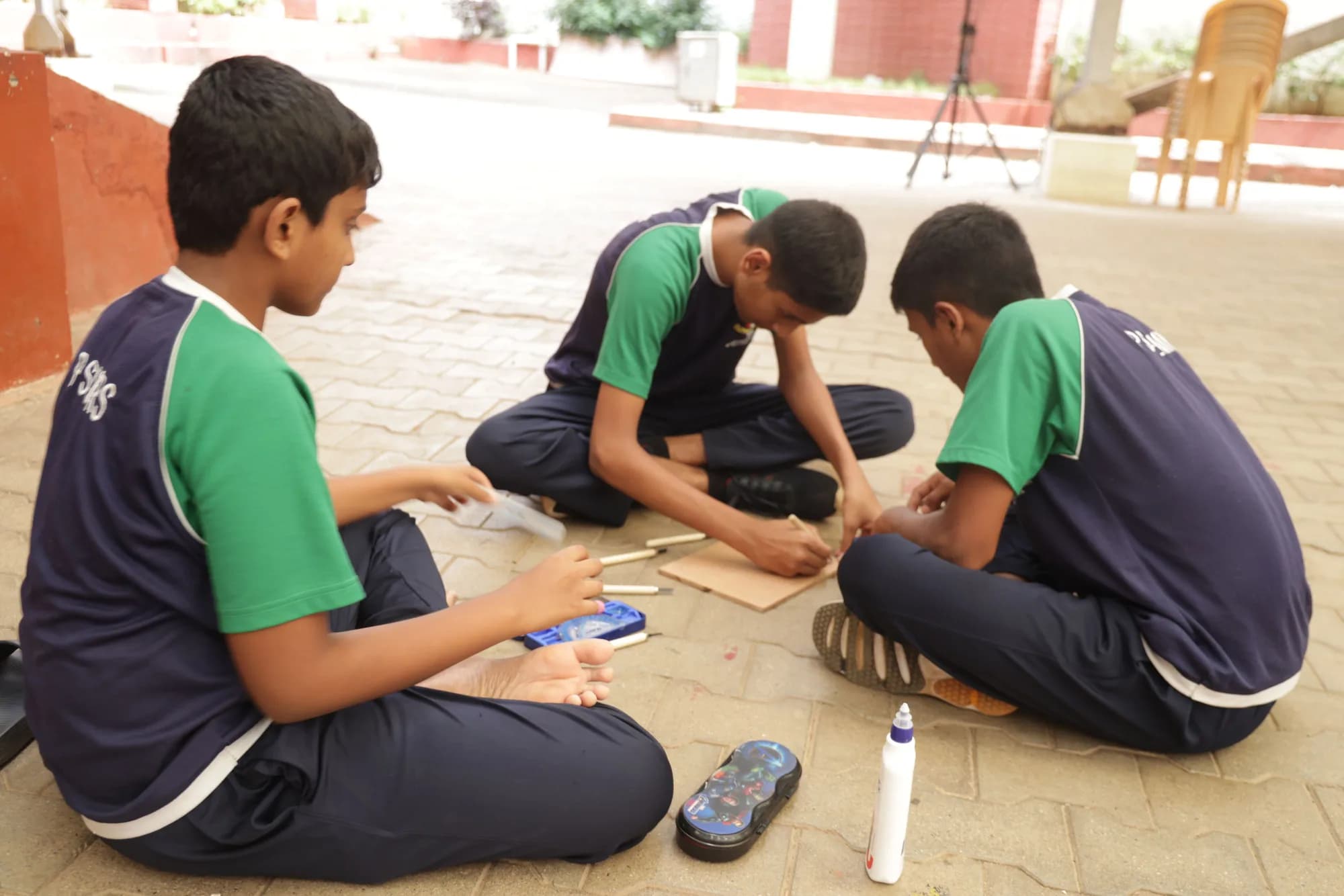 Three boys building a model from wooden sticks in a PSSEMR School courtyard