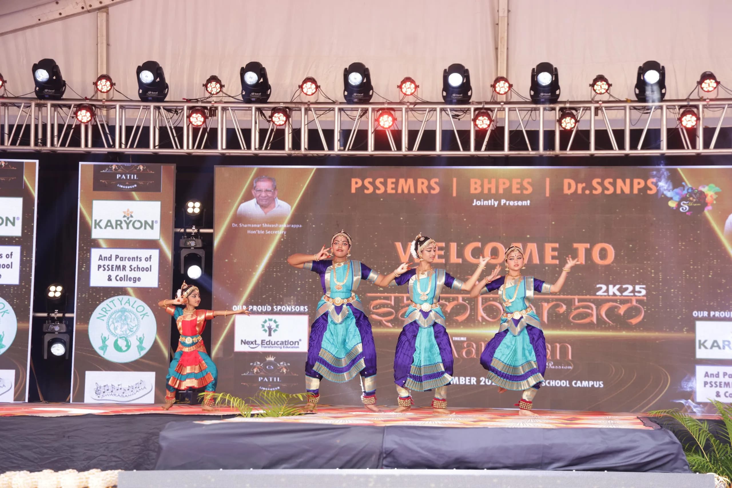 Four students in Bharatanatyam costume performing together on the main stage at Sambhram 2K25, under stage lighting.