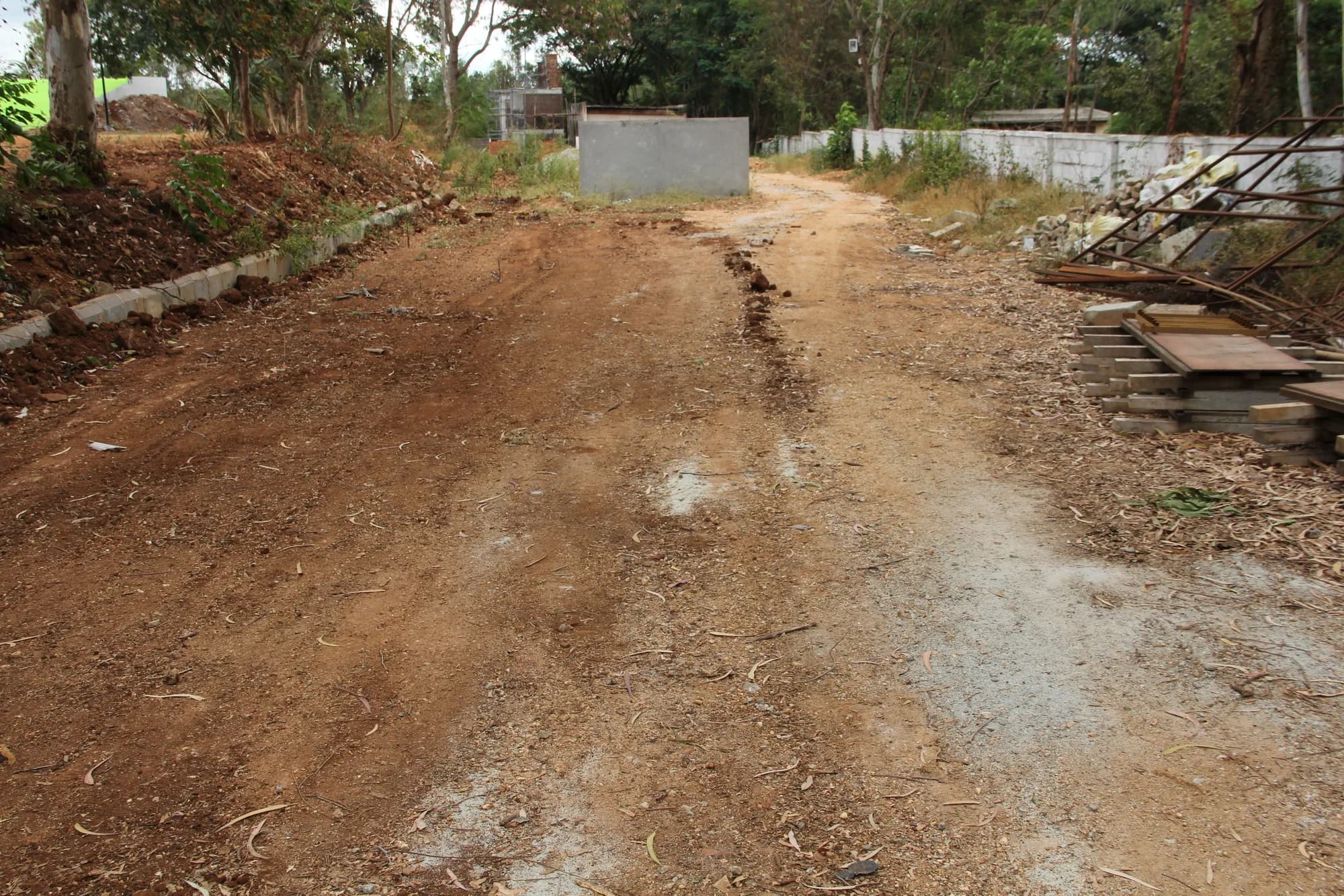 Undeveloped ground of bare red earth with building material stacked against a compound wall