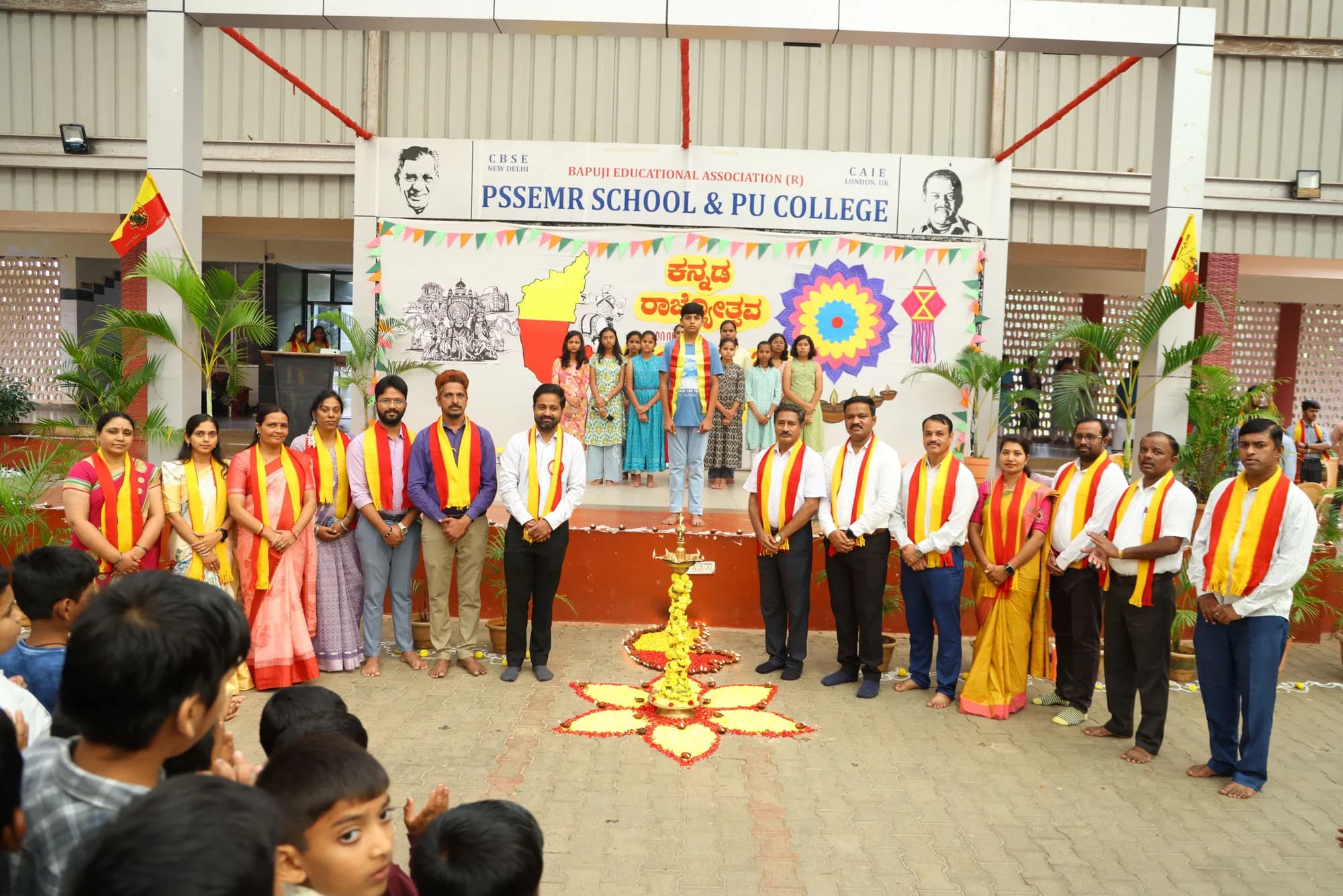 Staff in yellow and red shawls standing behind a lit ceremonial lamp