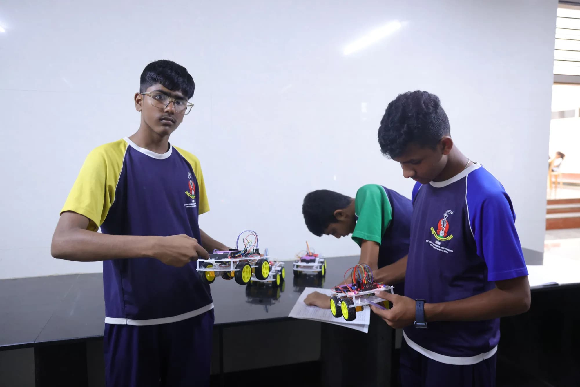Three students in house colours checking four-wheel robot builds at a laboratory bench