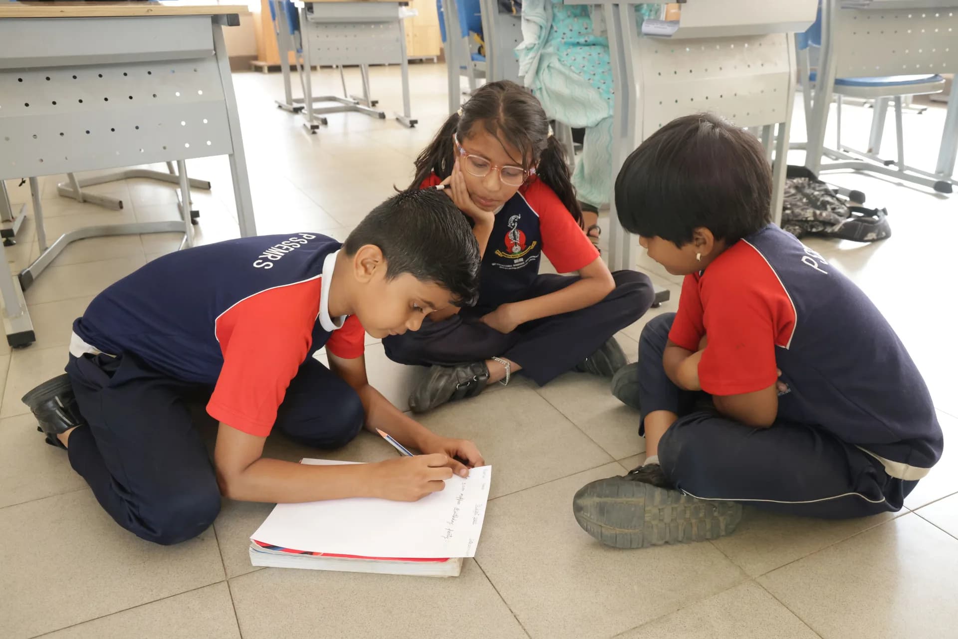 Children seated on the floor writing in Kannada on a sheet of paper