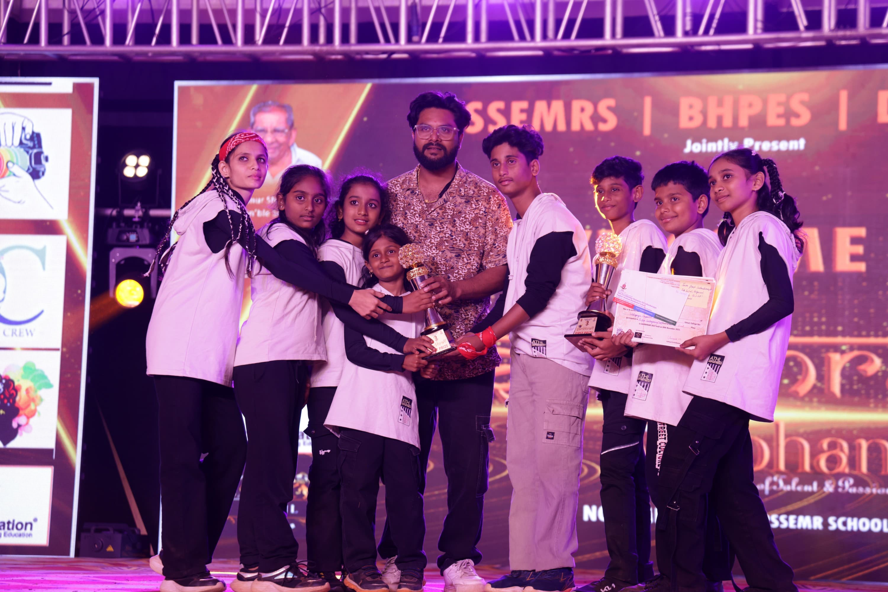 A winning team on the main stage at Sambhram 2K25 holding two first-prize trophies and their certificates, with their accompanying teacher standing among them.