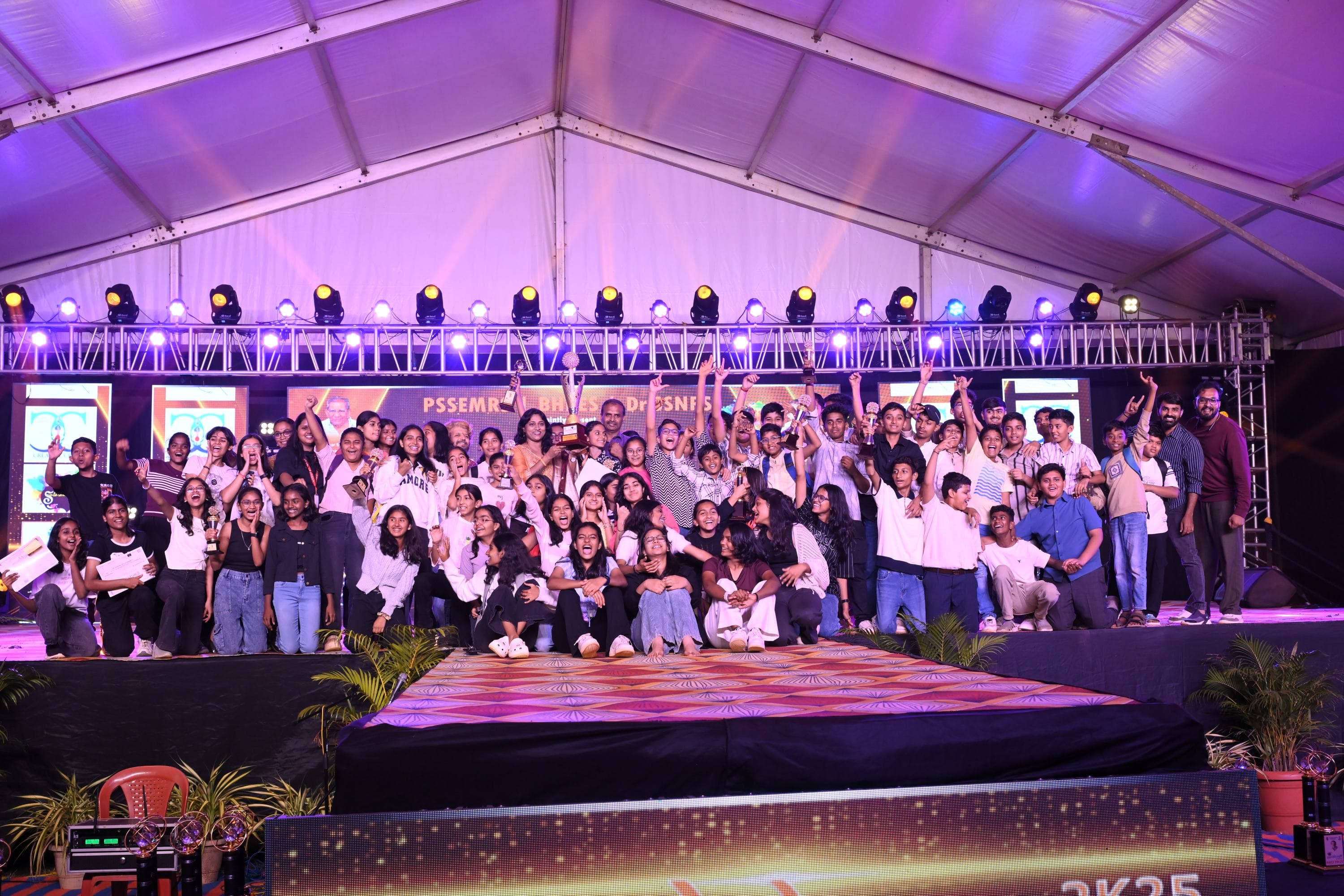 Around seventy students packed across the main stage at the close of Sambhram 2K25, trophies and certificates held up, with a teacher at the centre holding the overall trophy.